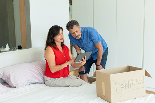 Couple packing donation box on bed in bedroom, woman wearing red blouse using tablet, man leaning