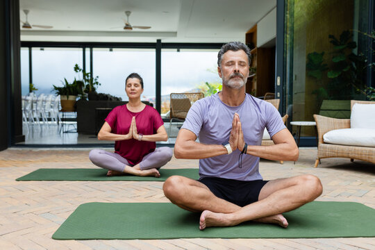 Diverse couple sitting meditating wearing sportswear on green yoga mats on patio by sliding doors