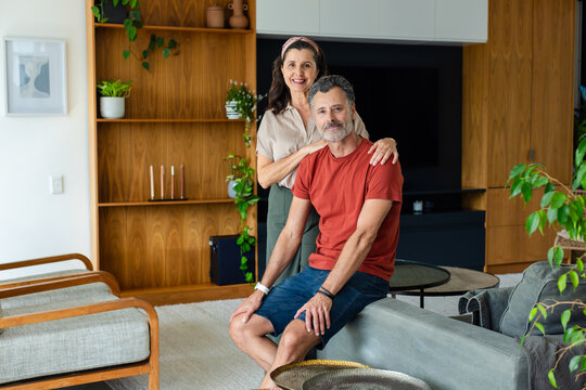 Diverse couple sitting on sofa arm in living room, man wearing rust T-shirt and wristwatch