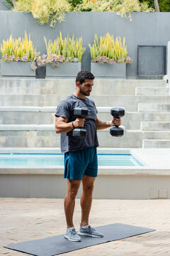 Adult man lifting hex dumbbells on exercise mat next to pool on patio in athletic gear