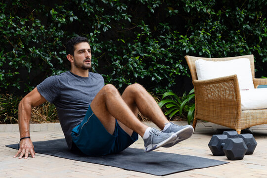 Man doing core exercise on black mat at patio wearing sportswear, hex dumbbells nearby