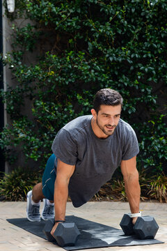 Man wearing gray tee holding hex dumbbells on black mat while performing high-plank on paved patio