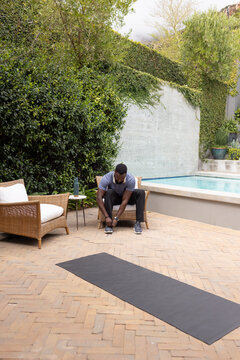 Man in sportswear sitting on armchair tying shoe on brick patio beside pool with black mat