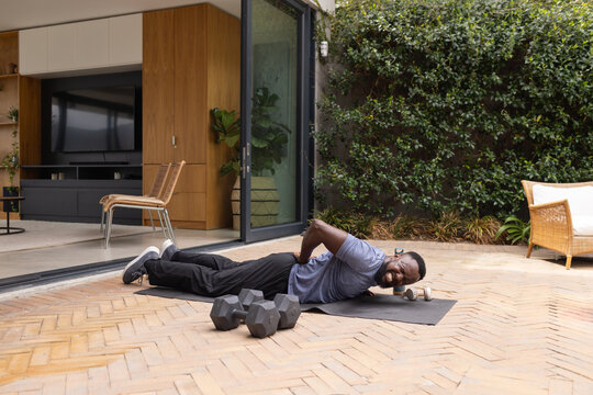 Adult African man lying on exercise mat clutching lower back in sportswear by dumbbells on patio
