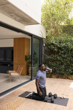 African American man kneeling on patio mat in sportswear drinking water with earbuds weights nearby