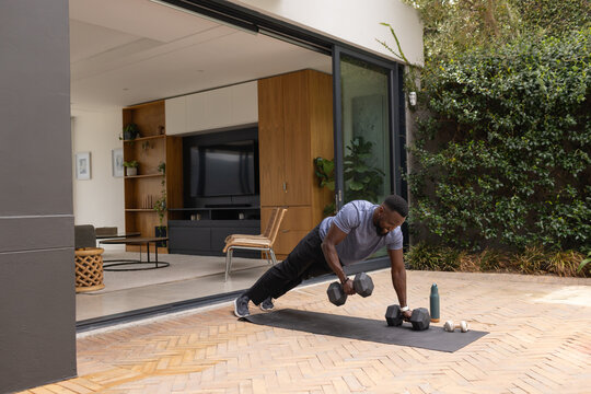 African American man wearing gray shirt exercising on patio mat holding hex dumbbells, copy space