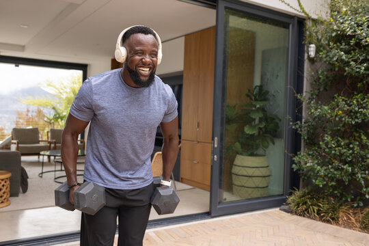Adult African American man lifting heavy hex dumbbell on patio by open sliding door wearing headset