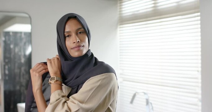 Female lifting headscarf, aligning and tucking, securing fit while checking mirror in bathroom