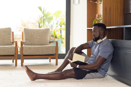 African American man sitting on floor in living room, in T-shirt, shorts, writing notebook with pen