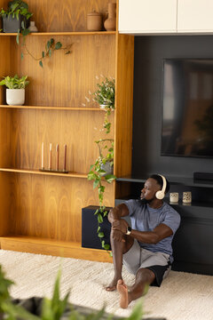 African American man sitting on rug at home wearing sportswear near mounted TV, copy space
