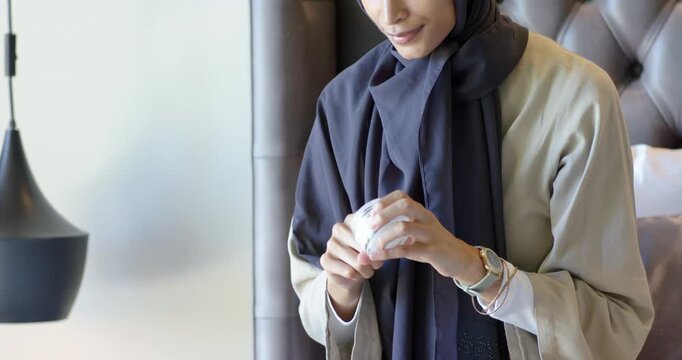 Adult woman on couch holding jar, opening lid, applying cream to hands for moisture, wearing hijab