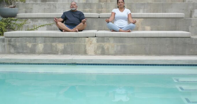 African American couple sitting cross-legged meditating by turquoise pool with planter reflections