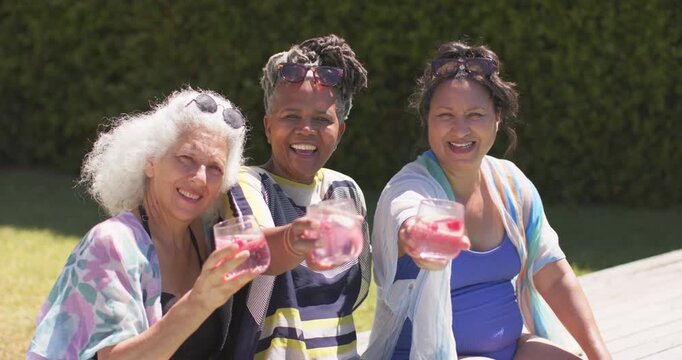 Female friends raising glasses with strawberry slices, toasting toward camera on pool deck