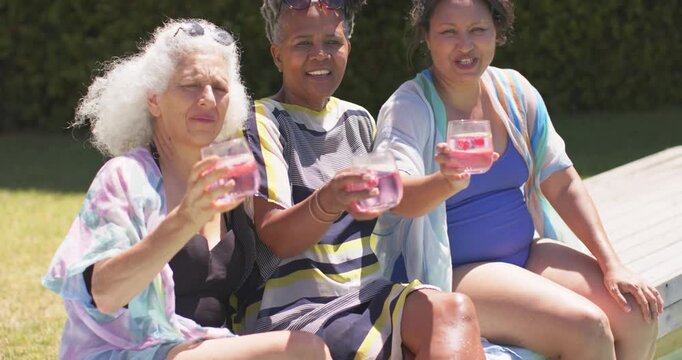 One woman is placing sunglasses, three women are raising stemless glasses and toasting poolside