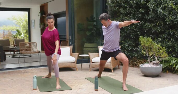 Couple starting yoga sequence by stepping into Warrior II on patio, improving balance on green mats