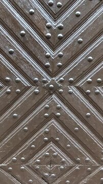 Vertical 4K footage with a steady camera move upwards, revealing a detailed dark brown antique wooden door. The surface features a striking chevron geometric pattern and rows of metal rivets.