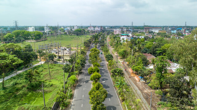 Urban aerial view of a widening road with a tree lined median, blending residential and industrial buildings, power lines and railway tracks into green suburban landscape