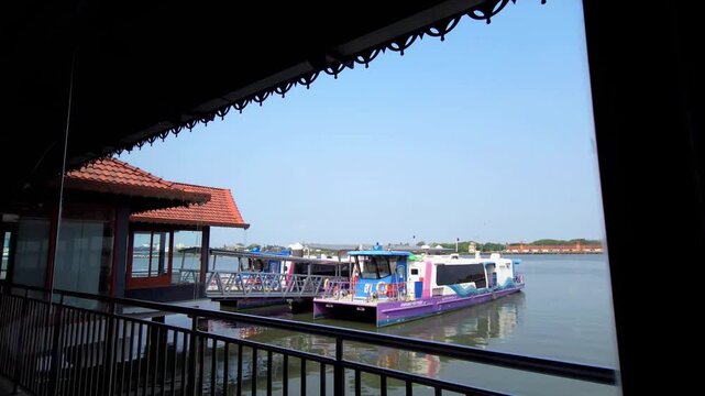 A view from a Kochi Water Metro terminal shows purple and white electric catamaran ferries docked on calm water under a clear sky. A traditional tiled-roof building and black railings frame the scene.