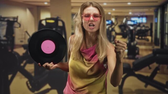 Blonde woman holding vinyl record, fist clenched and singing while wearing pink sunglasses in a gym building; retro workout fun.