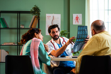 Obraz premium Indian doctor showing MRI scan results to retired Asian seniors across a table in a clinic.