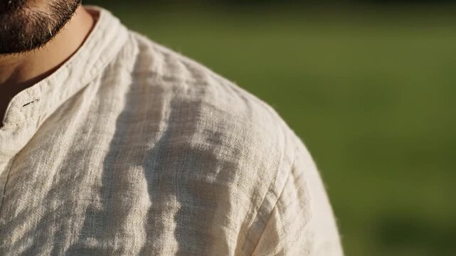 Close-up of a mans shoulder and beard, wearing a white linen shirt, with a blurred green background.
