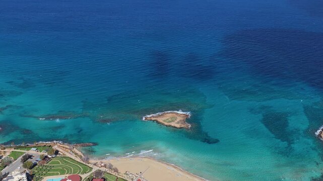 Aerial drone view of a beach with turquoise water and a small rocky island surrounded by shallow sea near the Cyprus coastline