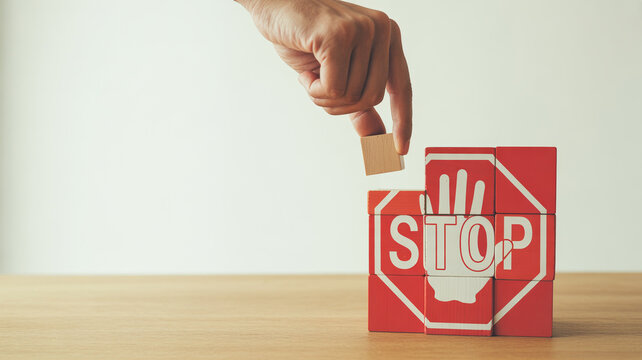 A human hand carefully places a small wooden block atop red cubes spelling the word STOP with a stop sign symbol on a clear light background creating 