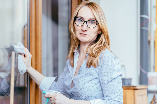 Smiling middle aged caucasian woman cleaning glass cabinet with spray at home