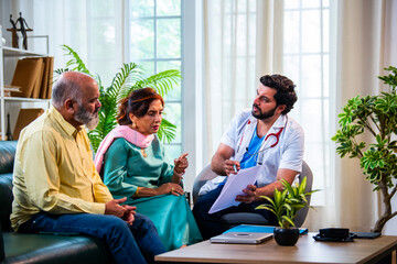 Obraz premium Indian doctor explaining a medical file to a senior Asian couple sitting on a clinic sofa.