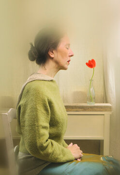 Woman sitting in front of a window in a meditative and romantic pose with a red tulip in her vase on a wooden shelf in the background