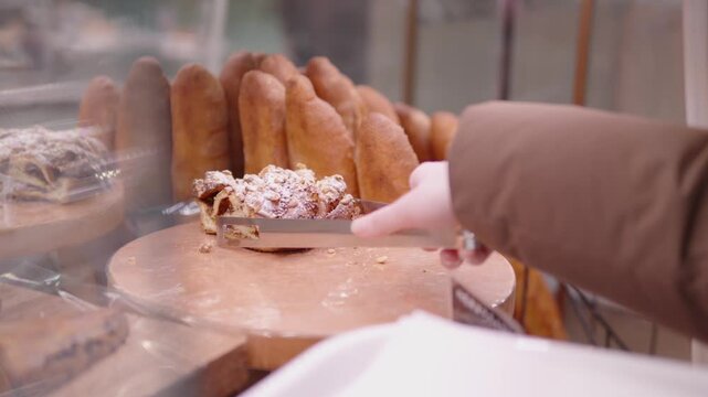Customer hand using serving tongs to pick nut topped sweet pastry from bakery display case, surrounded by fresh bread, baked goods for purchase, indulgence, highlights positive consumer experience
