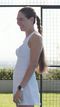 Vertical video: Smiling woman in white skort holding neon tennis ball on roof court reacting to cue