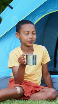 African American boy sitting by blue tent wearing yellow tee holding mug showing right knee bandage