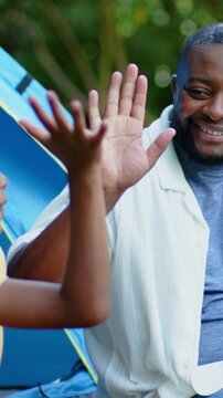 African American father and school-age child high-fiving while sitting in backyard with blue tent