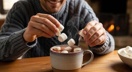 Cozy hot chocolate with marshmallows in cup by smiling man in sweater sitting at wooden table. Hot chocolate includes marshmallows being added to warm beverage, creating a comforting atmosphere.