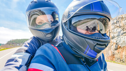 Two travelers wearing helmets on a motorcycle ride along a scenic road with rocky cliffs and blue sky in the background © JuliaDorian