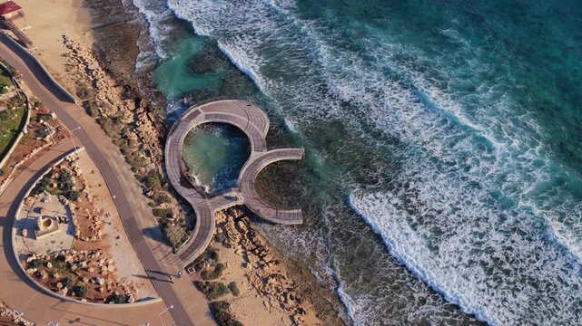 Aerial drone view of a coastal walkway and curved viewing platform along the shoreline in Ayia Napa, Cyprus, with waves crashing on rocks