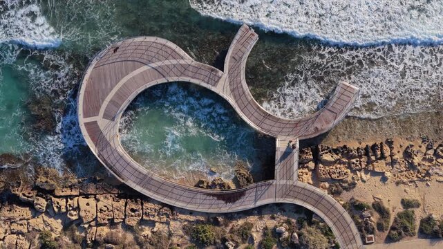 Aerial drone view of a unique curved wooden pier over the sea in Ayia Napa, Cyprus, surrounded by waves and rocky coastline
