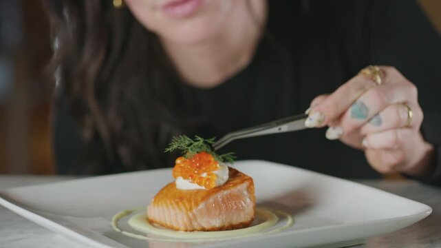 Chef places dill and salmon roe on a plated salmon dish using tweezers.