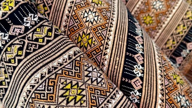 A panning close-up of traditional Thai hand-woven fabrics featuring intricate geometric patterns and earth-tone colors, showcasing the texture, weaving craftsmanship, and cultural details.
