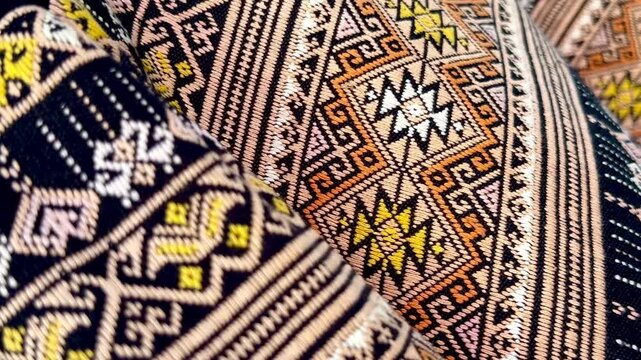 A panning close-up of traditional Thai hand-woven fabrics featuring intricate geometric patterns and earth-tone colors, showcasing the texture, weaving craftsmanship, and cultural details.