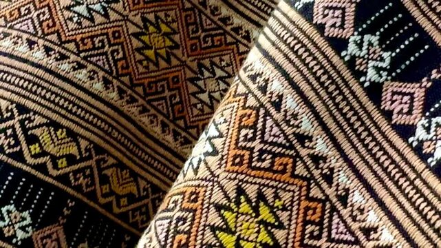 A panning close-up of traditional Thai hand-woven fabrics featuring intricate geometric patterns and earth-tone colors, showcasing the texture, weaving craftsmanship, and cultural details.