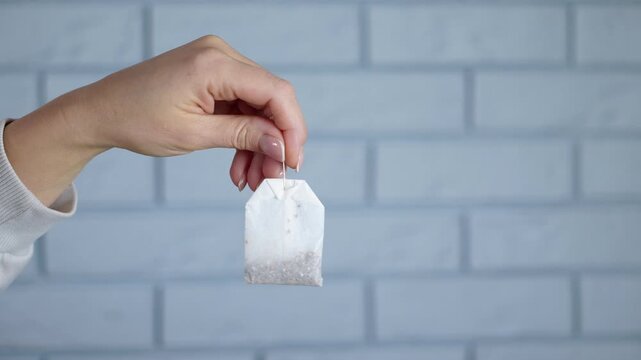 Woman holding a tea bag before brewing. Female hands holding a single paper tea bag with a string, preparing to brew a hot beverage against a soft-focus blue brick wall background
