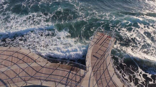 Aerial drone view of a unique curved wooden pier over the sea in Ayia Napa, Cyprus, surrounded by waves and rocky coastline