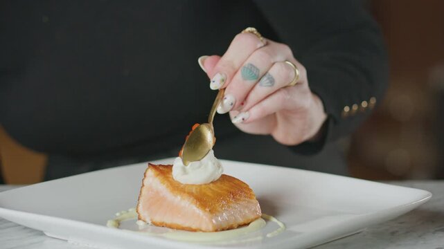 Chef garnishes a plated salmon dish with dill and caviar using tweezers.