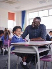 Fototapeta premium Black male teacher kneeling at eye level with a young student in a classroom, engaged in conversation with other students visible in the background