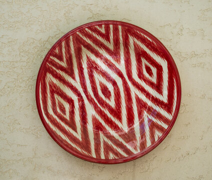 Traditional uzbek ceramic plate lagan with red atlas pattern hanging on stone wall. Rishtan Ceramics