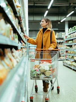 Woman in yellow coat standing in supermarket aisle, reaching for products on the shelf while pushing a shopping cart filled with groceries