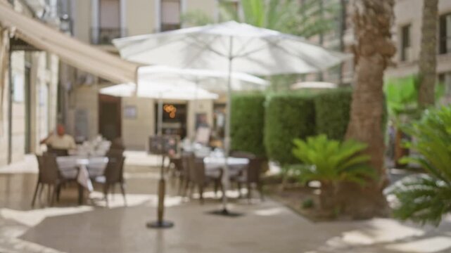 Blurry outdoor restaurant with white umbrellas and defocused chairs surrounded by greenery and buildings with a person sitting in the background
