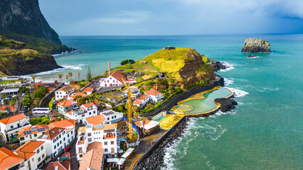 Aerial view of Porto da Cruz, Madeira, showcasing its unique black sand beach, natural volcanic pools, and charming coastal village against dramatic cliffs and the Atlantic Ocean. © Viktor
